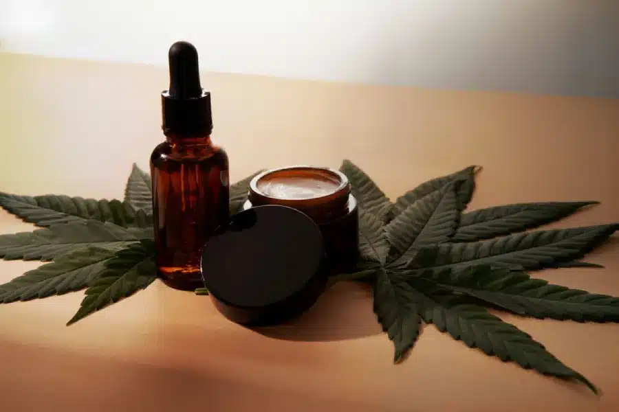 Cannabis-infused cosmetics with dropper bottle, cream jar and hemp leaves on neutral background