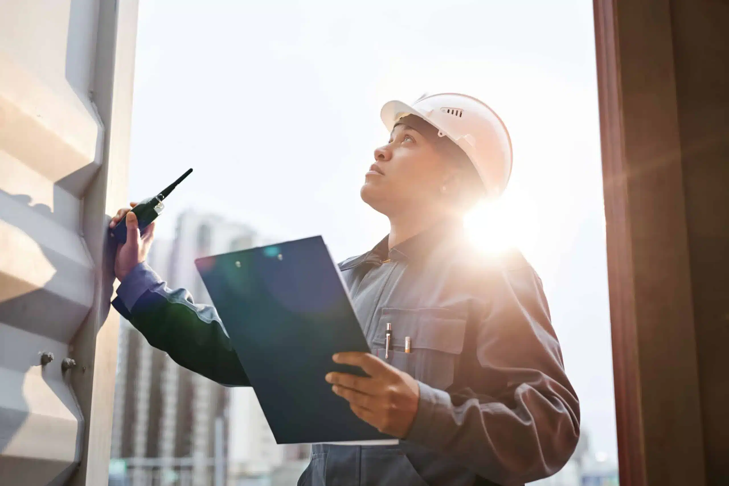 Construction worker in hard hat inspecting building with clipboard and radio in bright sunlight