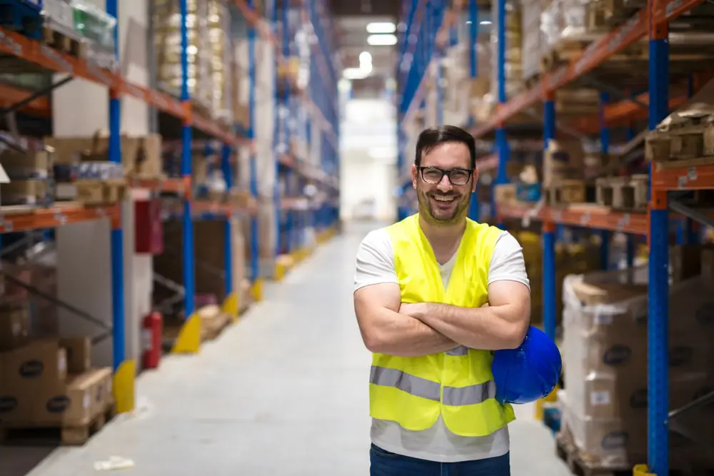 Smiling warehouse worker in safety vest standing in distribution center aisle