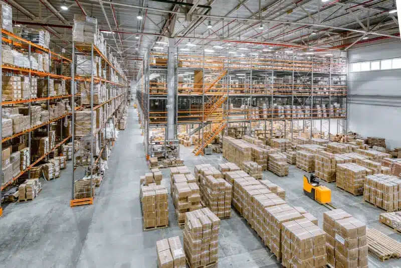 Large modern warehouse interior with stacked boxes, industrial shelving, and yellow forklift