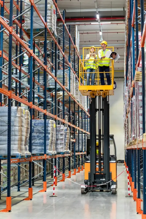 Two warehouse workers in safety gear operating yellow lift machine between tall storage racks