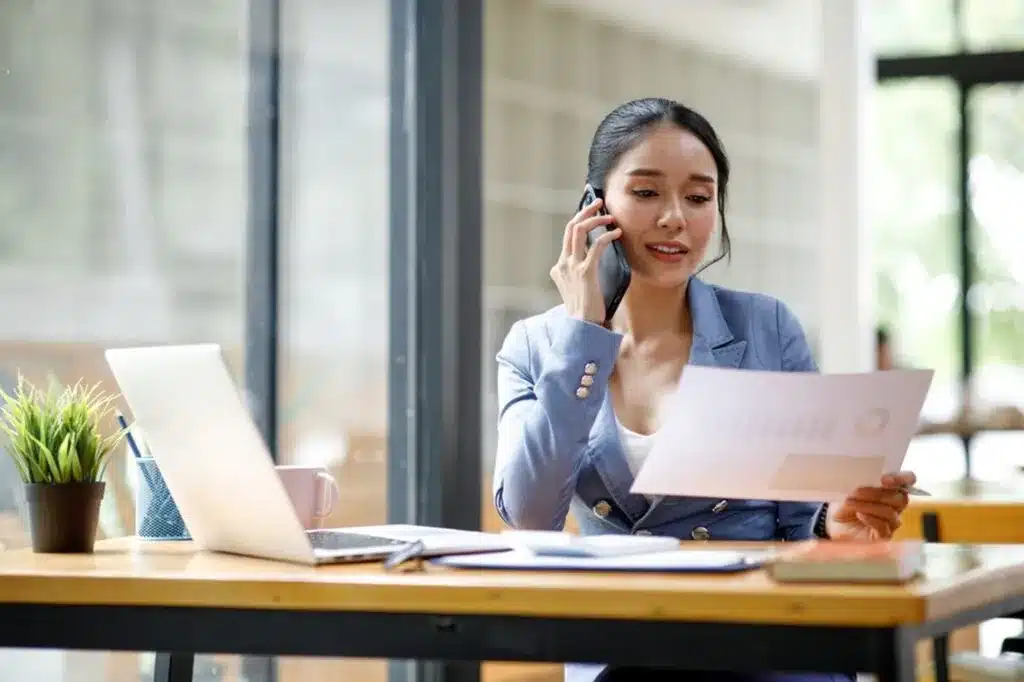 Professional businesswoman making phone call while reviewing financial documents at office desk