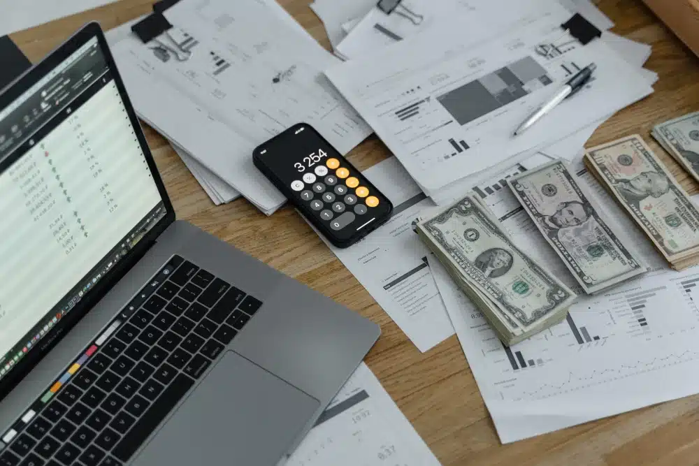 laptop, report print outs, calculator and stacks of cash on desk