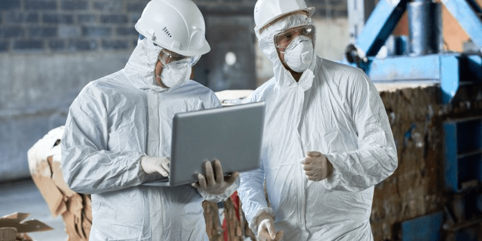 two people in protective suits, masks, hard hats and safety glasses. One typing on a laptop