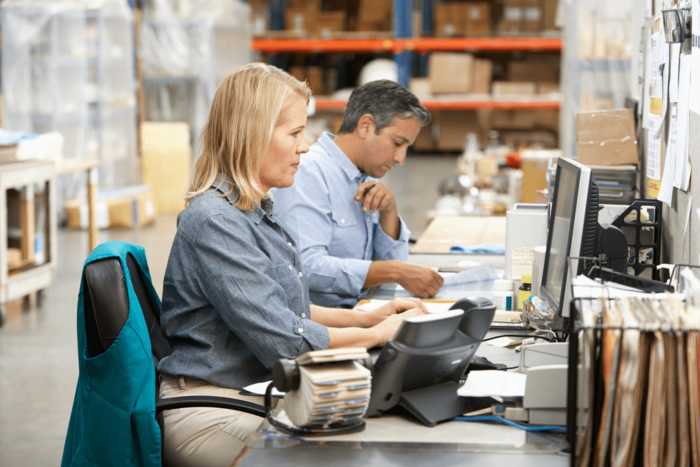 2 warehouse workers at a desk one looking at a computer and typing and another one looking at a book