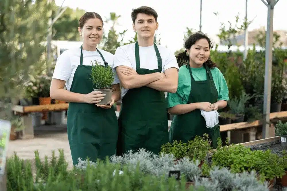 Three nursery employees in green aprons standing among potted plants and herbs